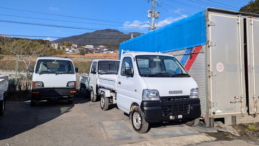 Three kei trucks sit next to a container awaiting shipping.